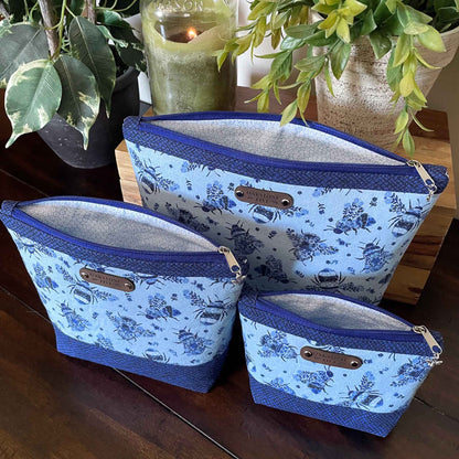Set of 3 blue floral bumble bee patterned zipper pouches, sitting open to show the interior, on a wooden table with a candle and plants.