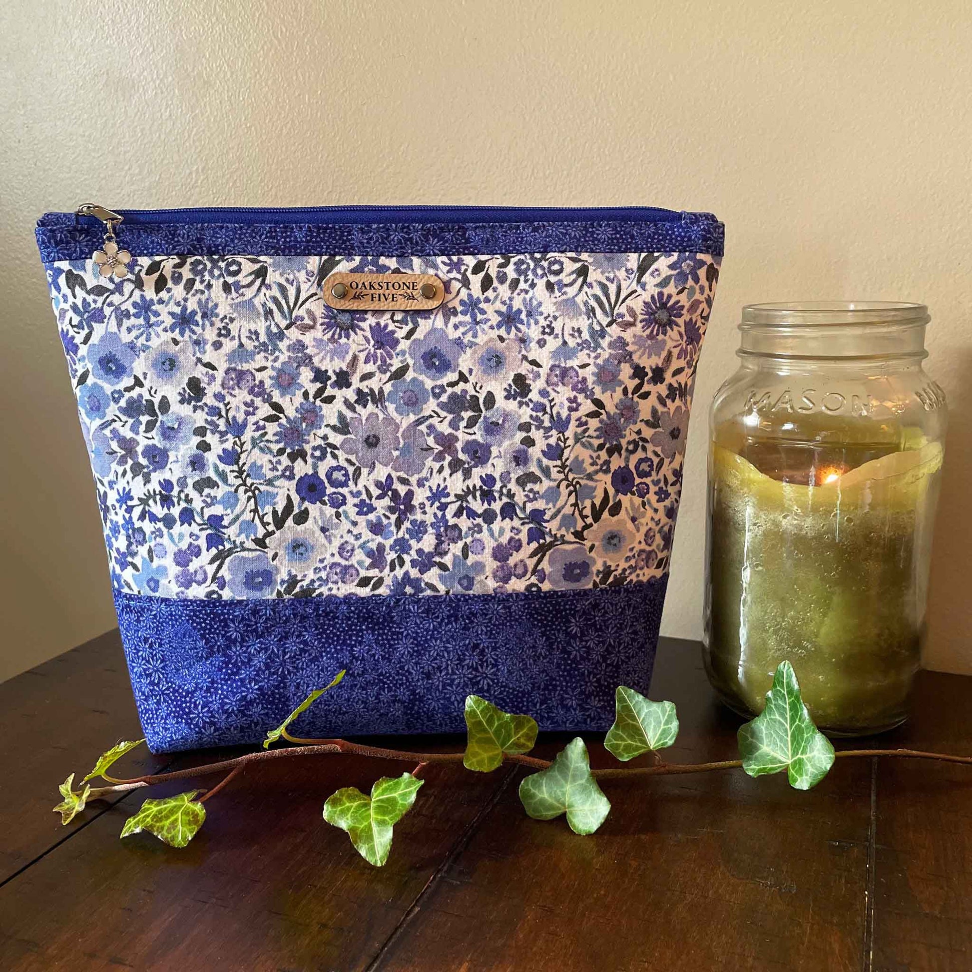 Size large, blue floral-patterned zipper pouch next to a lit candle in a jar on a wooden table with ivy leaves.