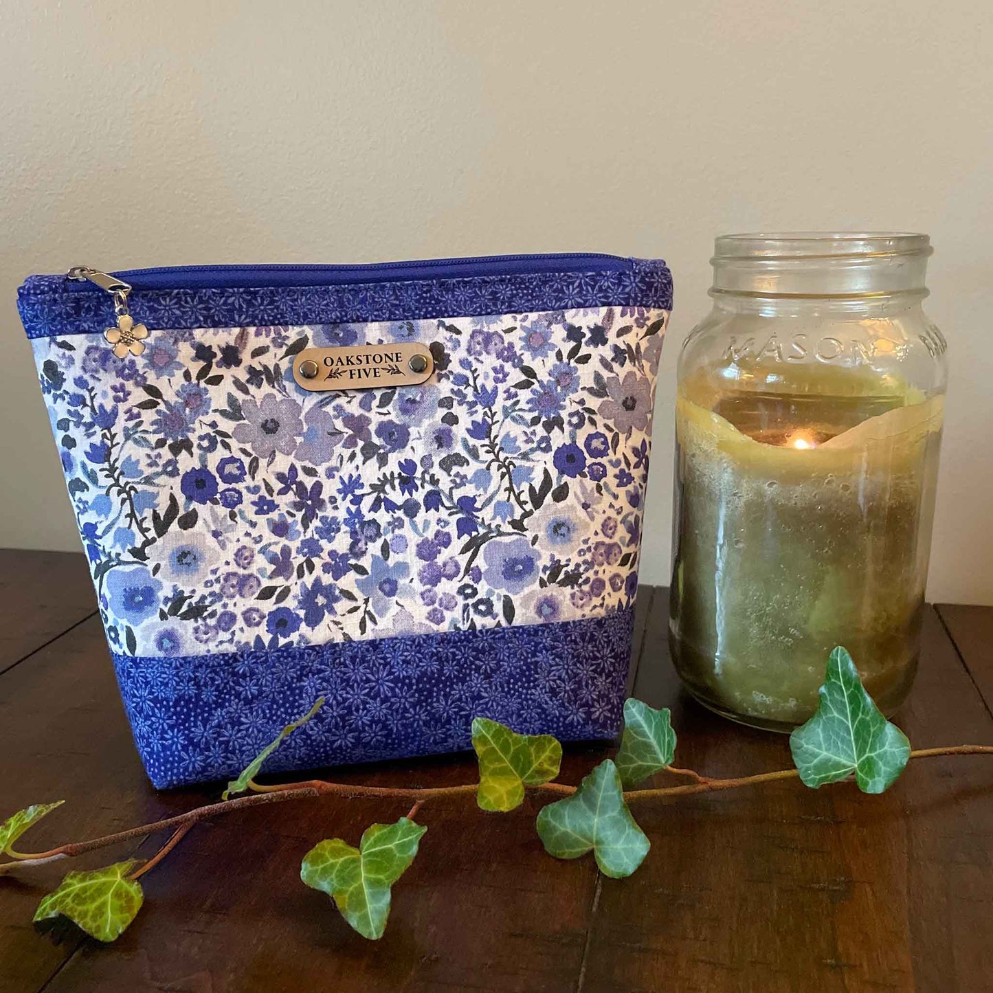 Size medium, blue floral-patterned zipper pouch next to a lit candle in a jar on a wooden table with ivy leaves.