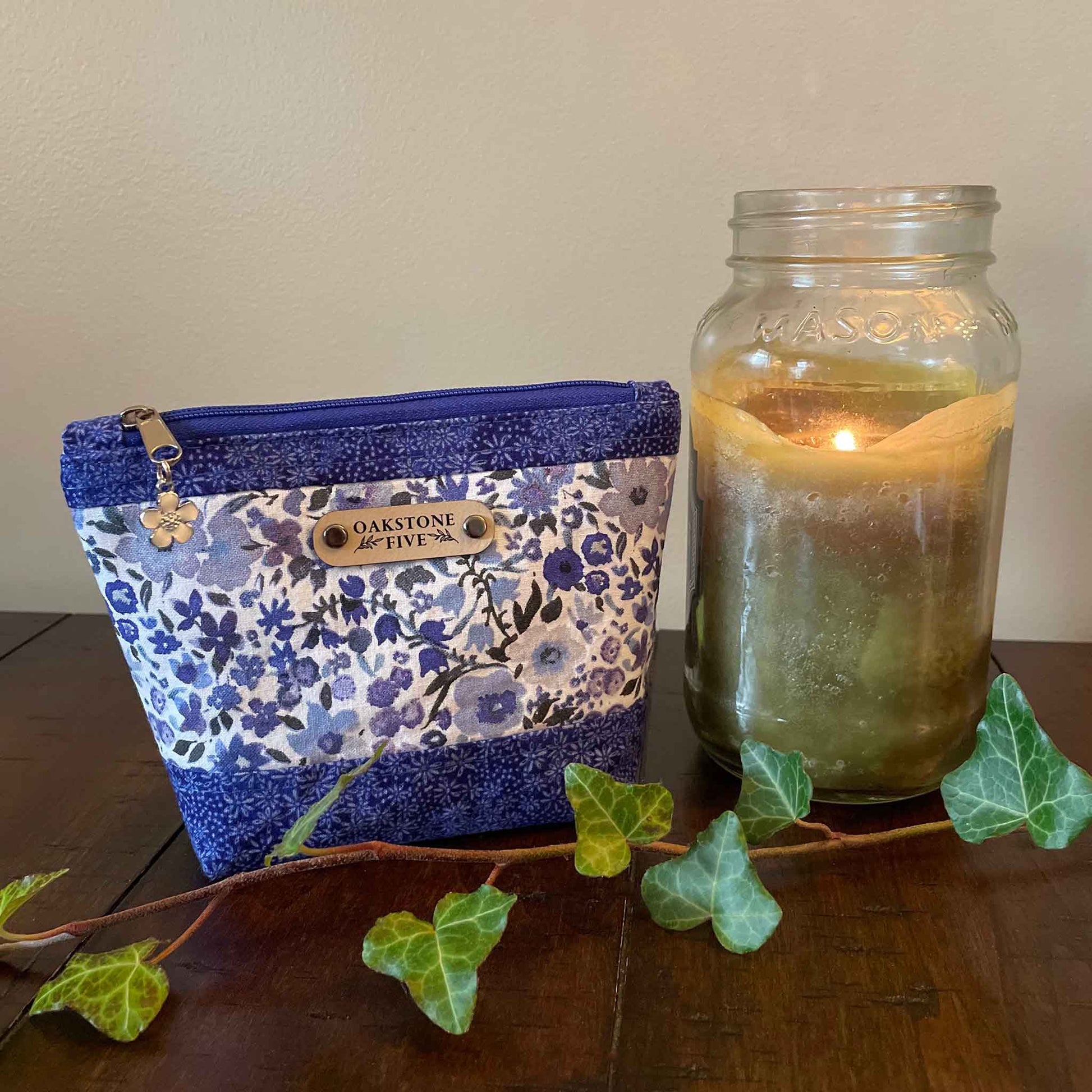 Size small, blue floral-patterned zipper pouch next to a lit candle in a jar on a wooden surface with ivy leaves.