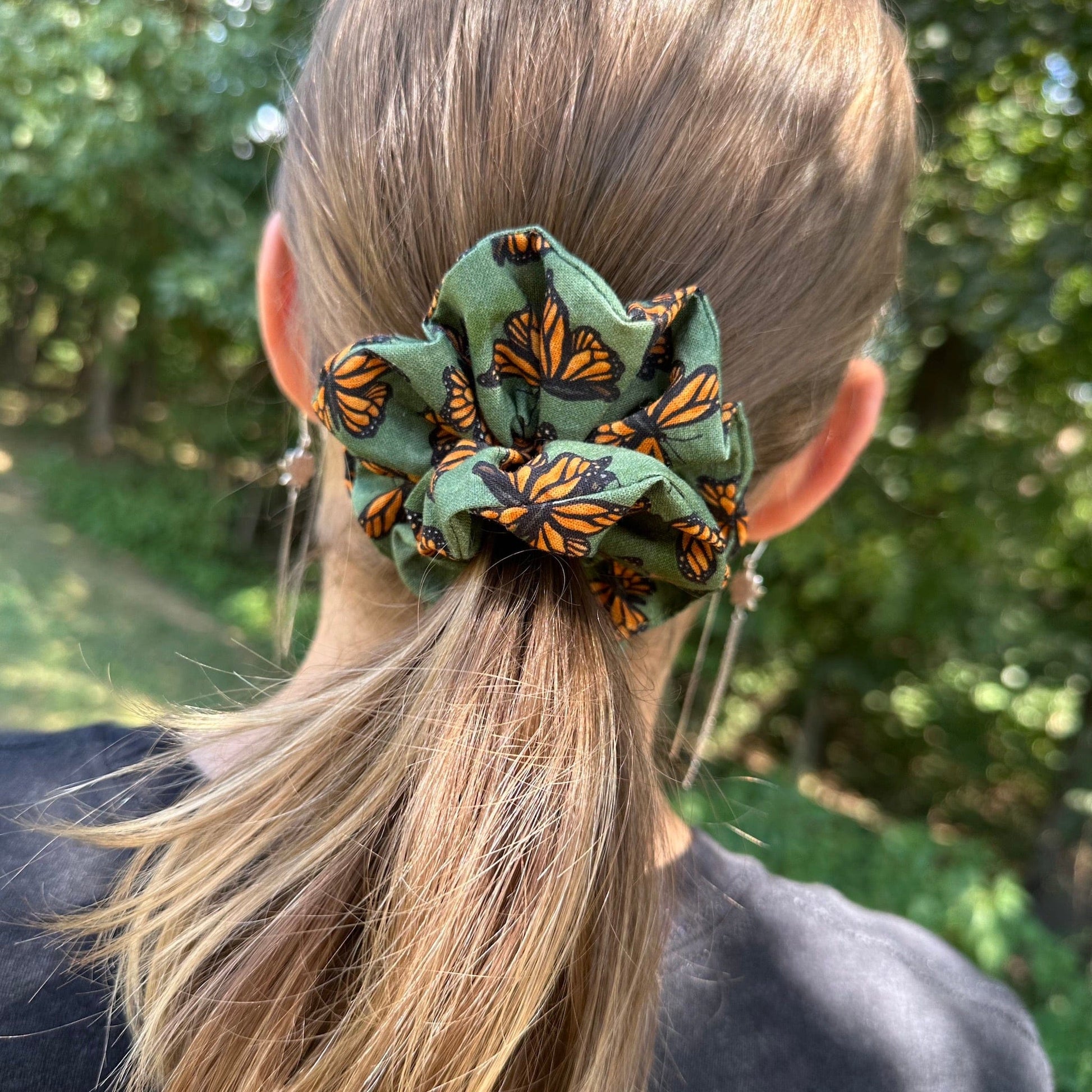 A girl standing in the woods, wearing a green, monarch butterfly scrunchie in her hair.