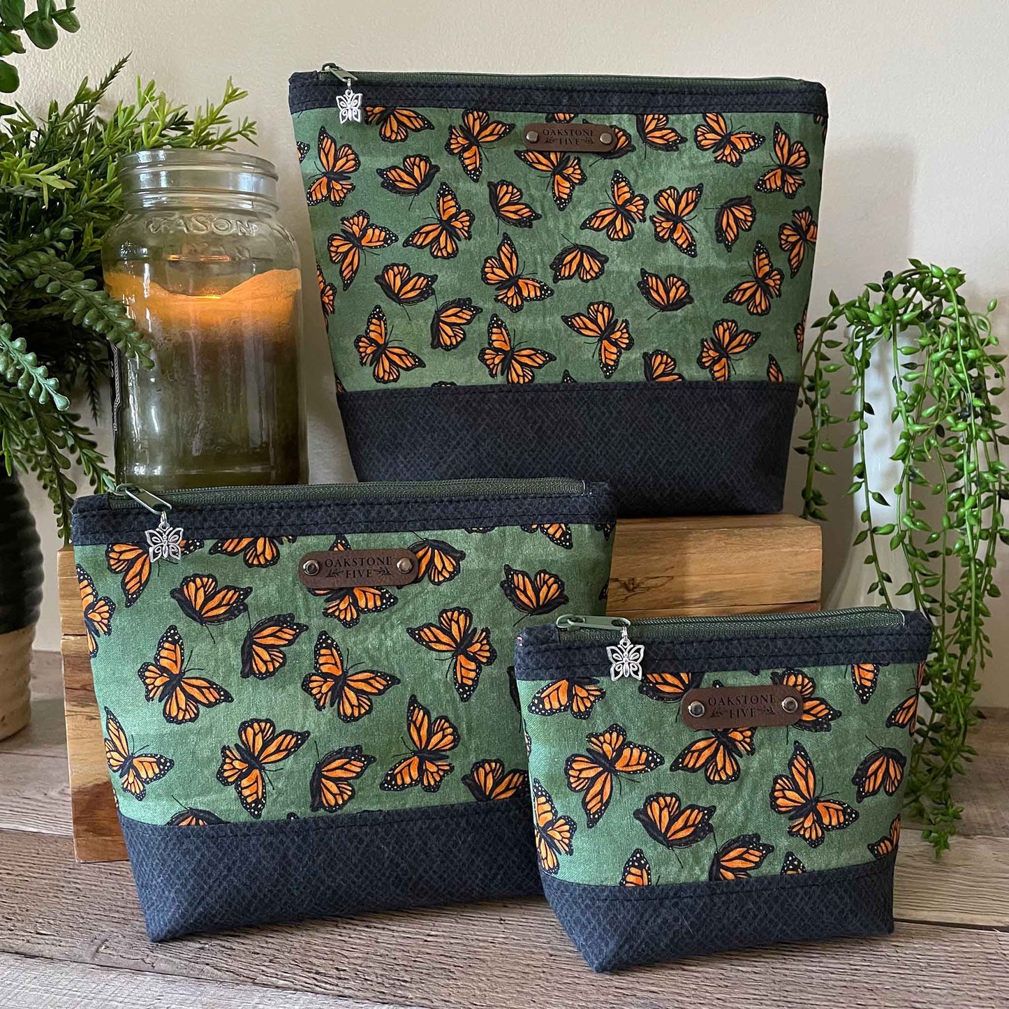 Three green zipper pouches with orange monarch butterfly pattern on a wooden surface with plants and a candle in the background.