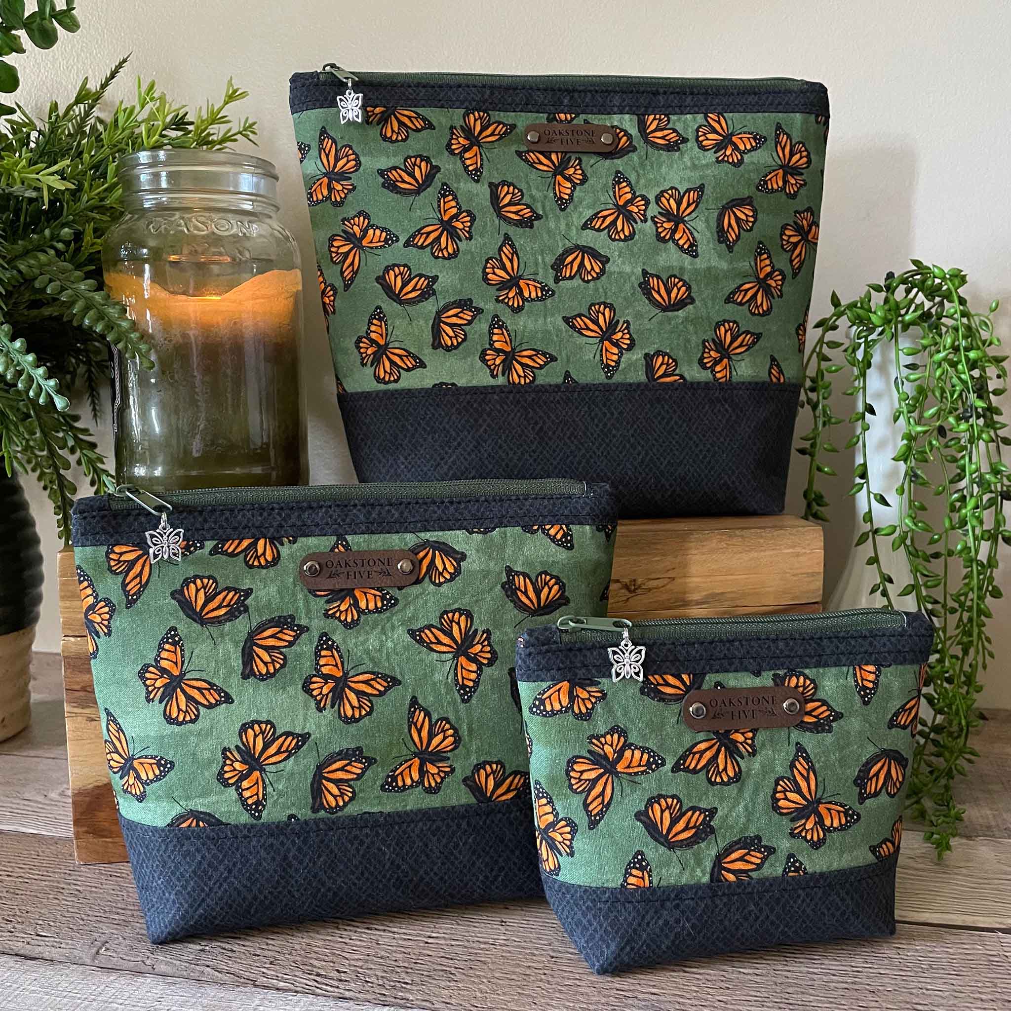 Three green zipper pouches with orange monarch butterfly pattern on a wooden surface with plants and a candle in the background.