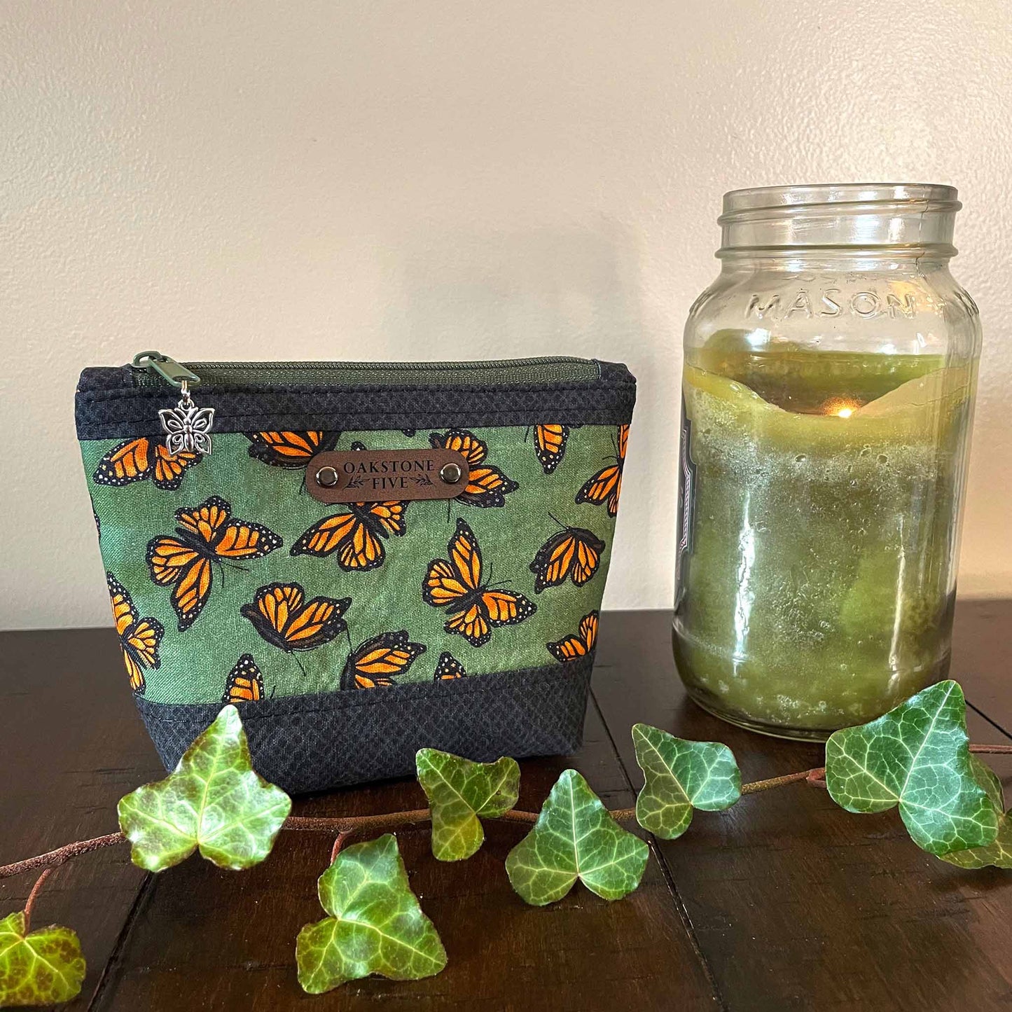 Small zipper pouch with monarch butterfly pattern next to a mason jar candle on a wooden surface.