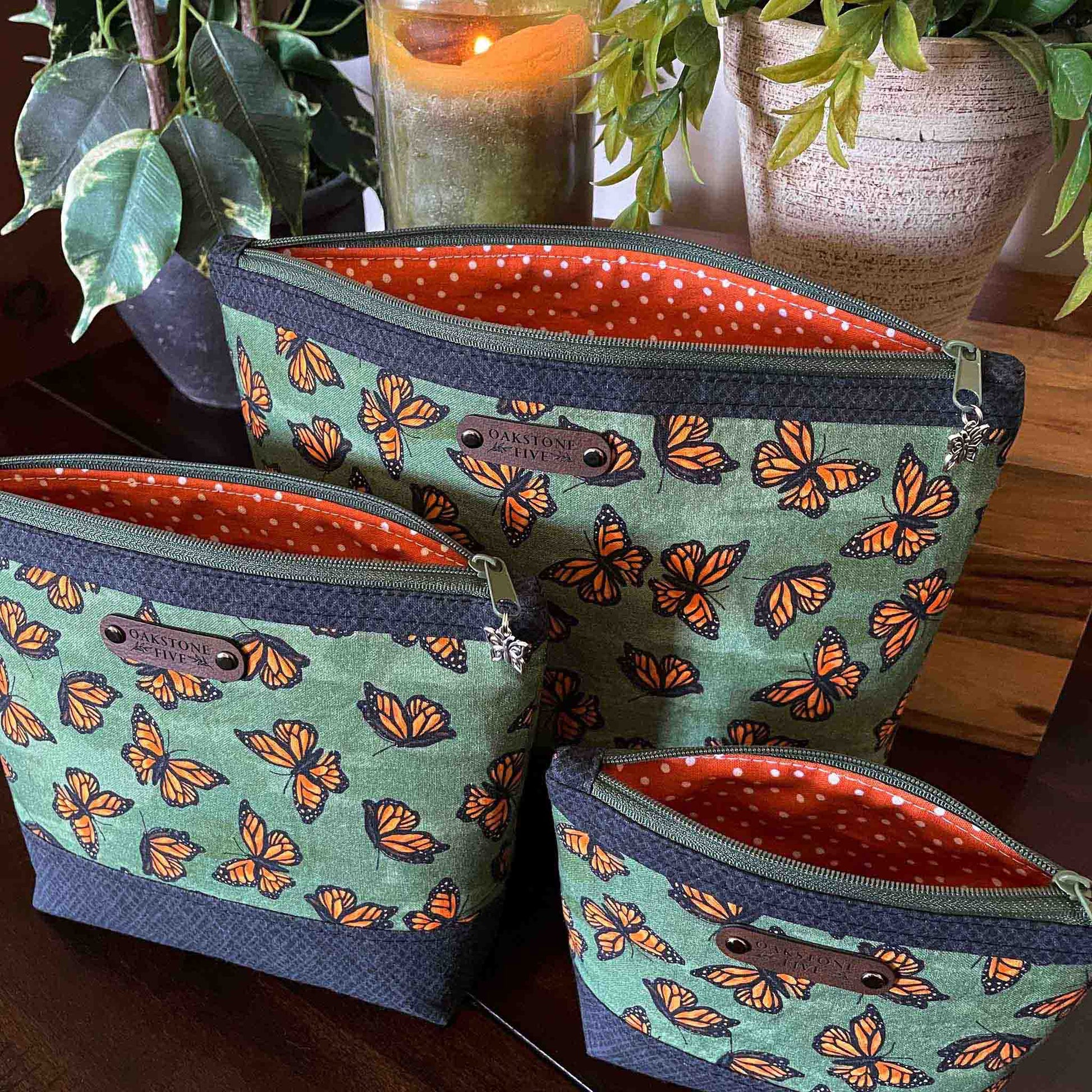 Three green zipper pouches with a monarch butterfly pattern and orange and white polka dot interior on a wooden surface with plants and a candle