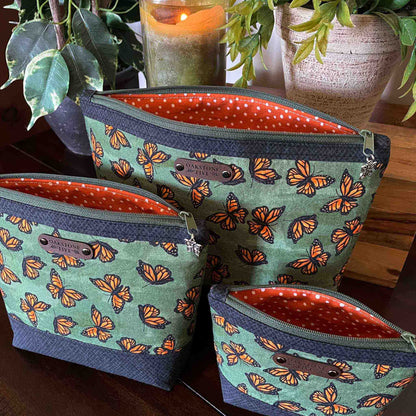 Three green zipper pouches with a monarch butterfly pattern and orange and white polka dot interior on a wooden surface with plants and a candle