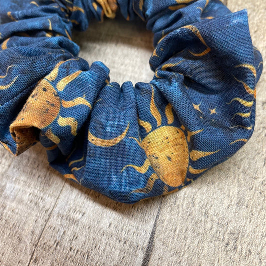 An up-close image of blue, handmade scrunchie with a sun, moon, and stars pattern laying on a wood background.