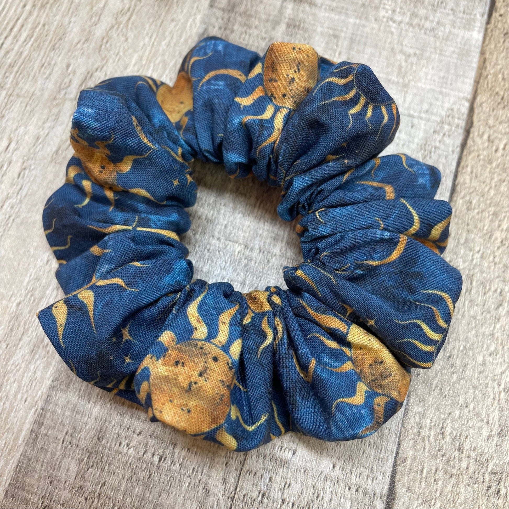 A blue, handmade scrunchie with a sun, moon, and stars pattern laying on a wood background.