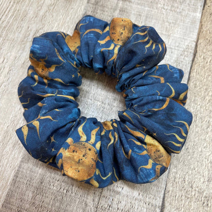 A blue, handmade scrunchie with a sun, moon, and stars pattern laying on a wood background.