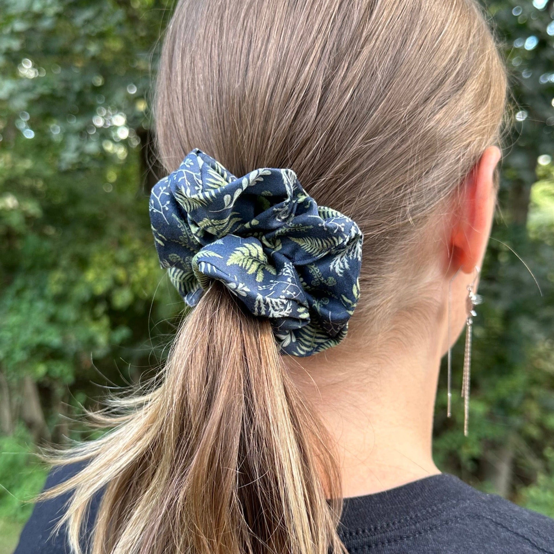 A photo of a navy ferns hair scrunchie being worn in a girl's ponytail, with a nature background.