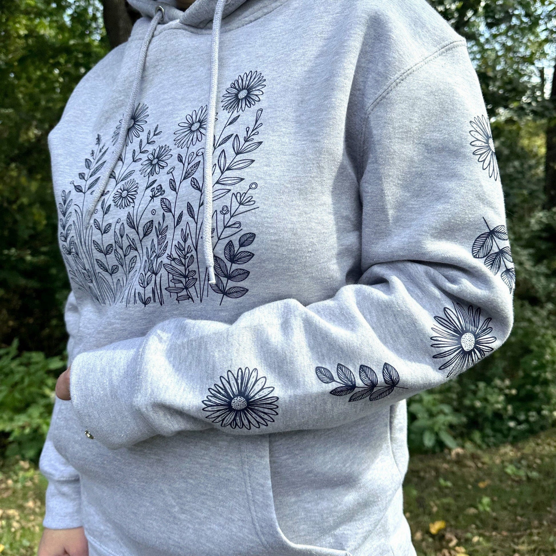 A woman standing outdoors wearing a fun, light grey hoodie that navy wildflowers on the front and on both sleeves.