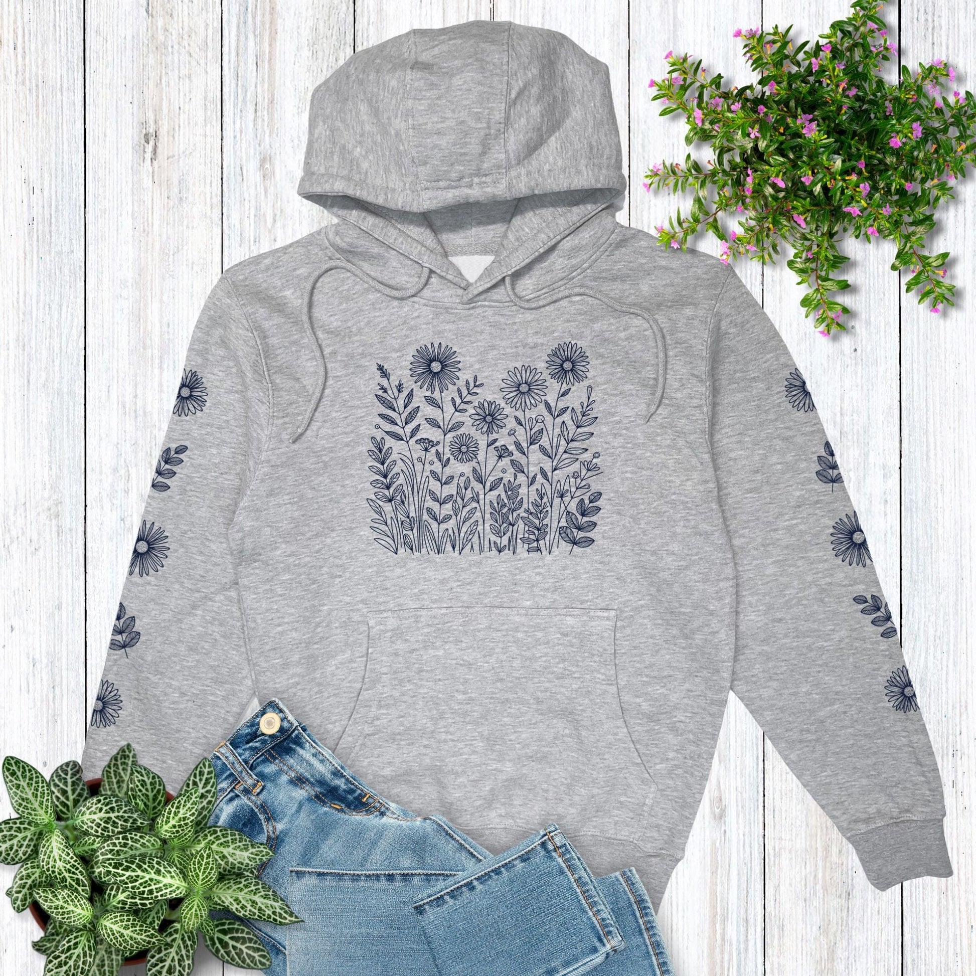 Gray hoodie with floral design on the front and on both sleeves, laying on a wooden surface with plants around