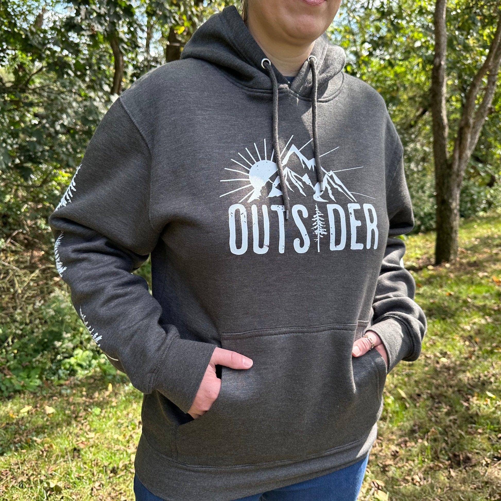 Woman standing in the woods, wearing a gray hoodie that says "Outsider" on the front with a sun and mountain scene, and has trees going down the sleeves.