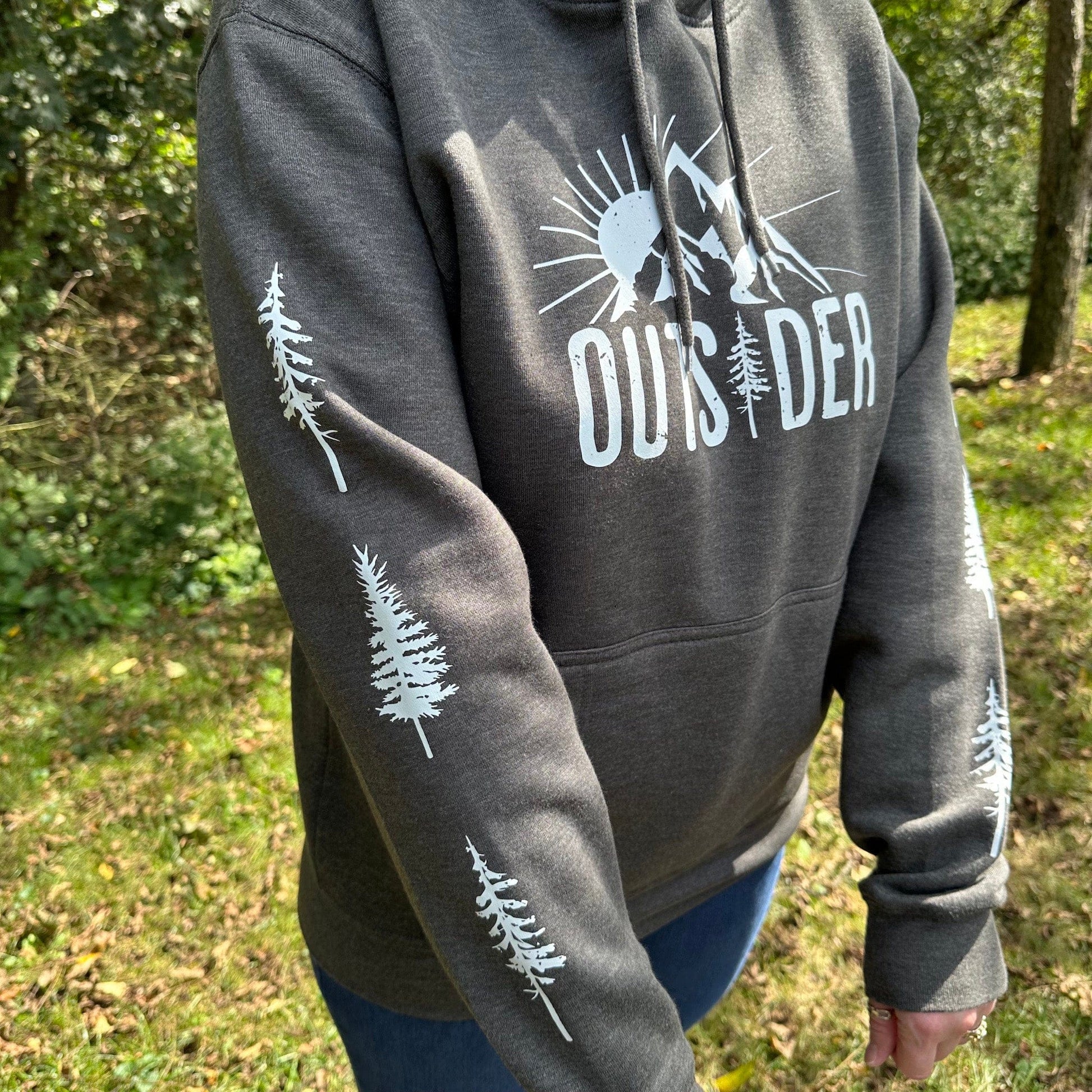 Person standing in the woods, wearing a gray hoodie that says "Outsider" on the front with a sun and mountain scene, and has trees going down the sleeves.