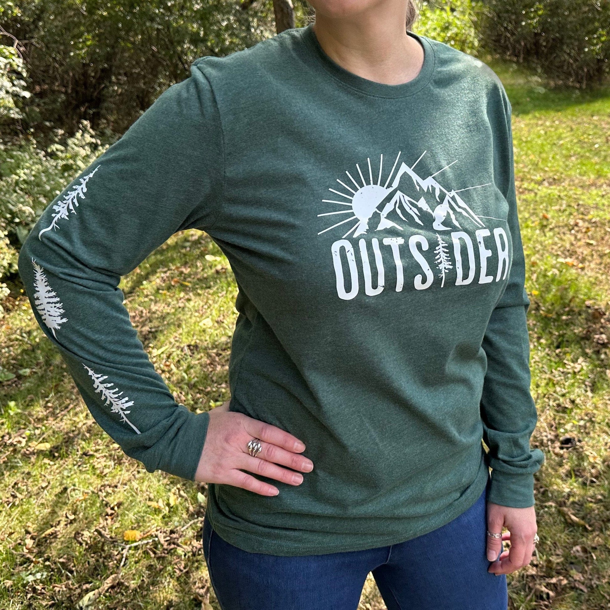 Woman standing outdoors in the woods, wearing a green long sleeve shirt that says "Outsider" on the front and has white trees down the sleeves.