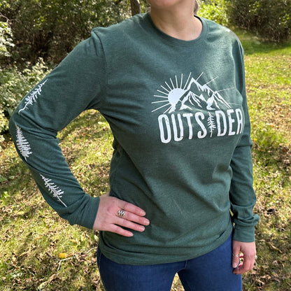 Woman standing outdoors in the woods, wearing a green long sleeve shirt that says "Outsider" on the front and has white trees down the sleeves.
