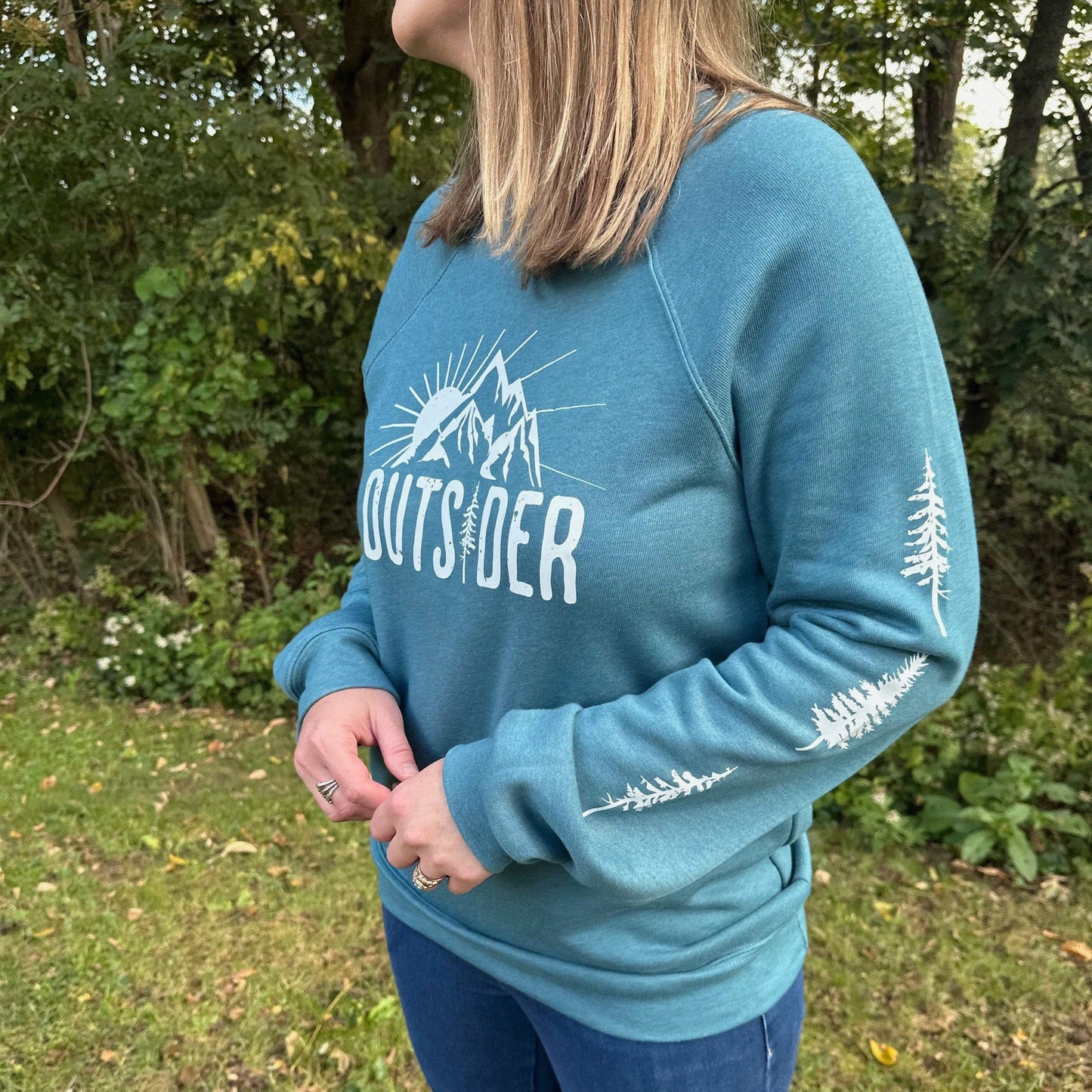 Woman standing outside among trees, wearing a teal nature sweatshirt with an 'OUTSIDER'  design on the front and trees down both sleeves.