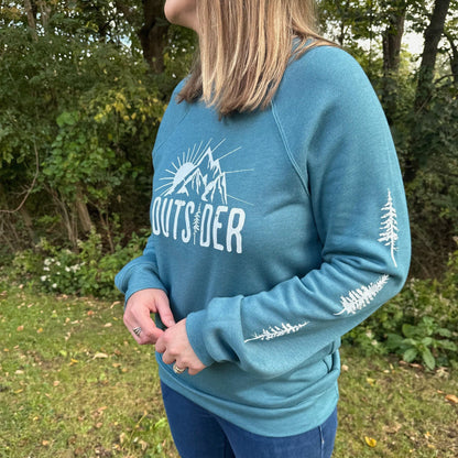 Woman standing outside among trees, wearing a teal nature sweatshirt with an 'OUTSIDER'  design on the front and trees down both sleeves.