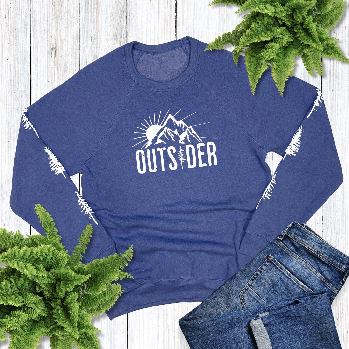 Blue sweatshirt that says, "Outsider" and has trees down each sleeve, laying on a wood background with jeans and plants.