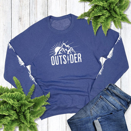 Blue sweatshirt that says, "Outsider" and has trees down each sleeve, laying on a wood background with jeans and plants.