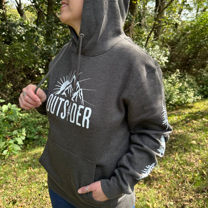 Woman wearing a gray hoodie that says "Outsider" on the front and has a sun and mountain scene, and trees going down the sleeves.