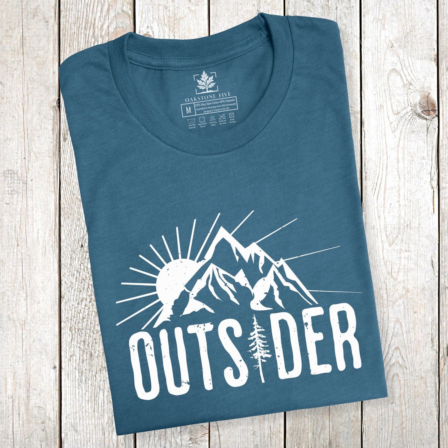 A folded teal-colored mountain shirt for nature lovers that says "Outsider" and shows mountain peaks with sunshine.