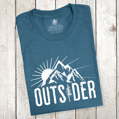 A folded teal-colored mountain shirt for nature lovers that says "Outsider" and shows mountain peaks with sunshine.