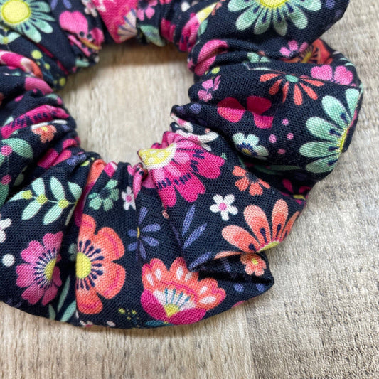 A close-up of a pink, green floral patterned scrunchie on a wood background