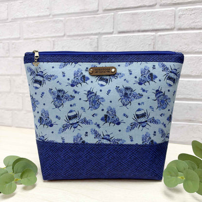 A large, blue zipper pouch with a pollinator bee pattern accented with a royal blue fabric, sitting on a white surface with leaves, against a white brick wall.