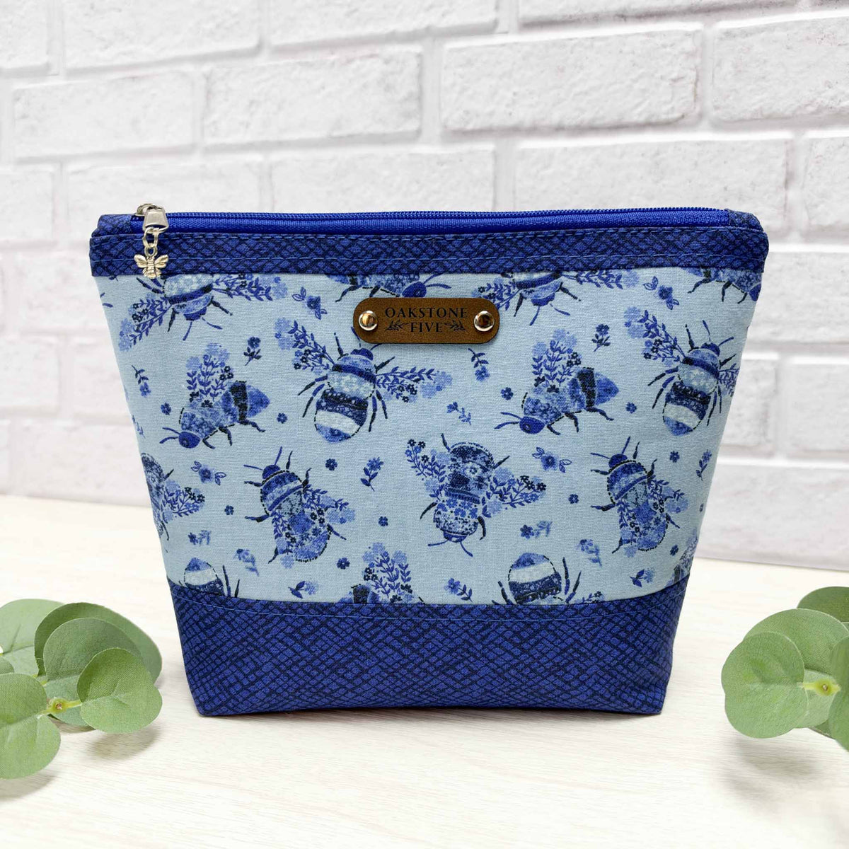 A blue floral bumblebee patterned zipper pouch with royal blue accents, a silver bee charm on the the zipper and a faux leather "Oakstone Five" brand tag, sitting on a white surface with leaves in front of a white brick background.