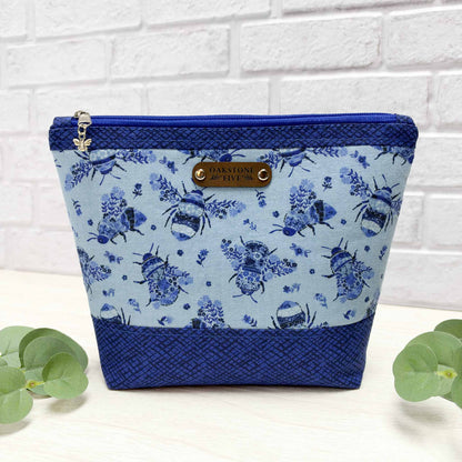 A blue floral bumblebee patterned zipper pouch with royal blue accents, a silver bee charm on the the zipper and a faux leather "Oakstone Five" brand tag, sitting on a white surface with leaves in front of a white brick background.