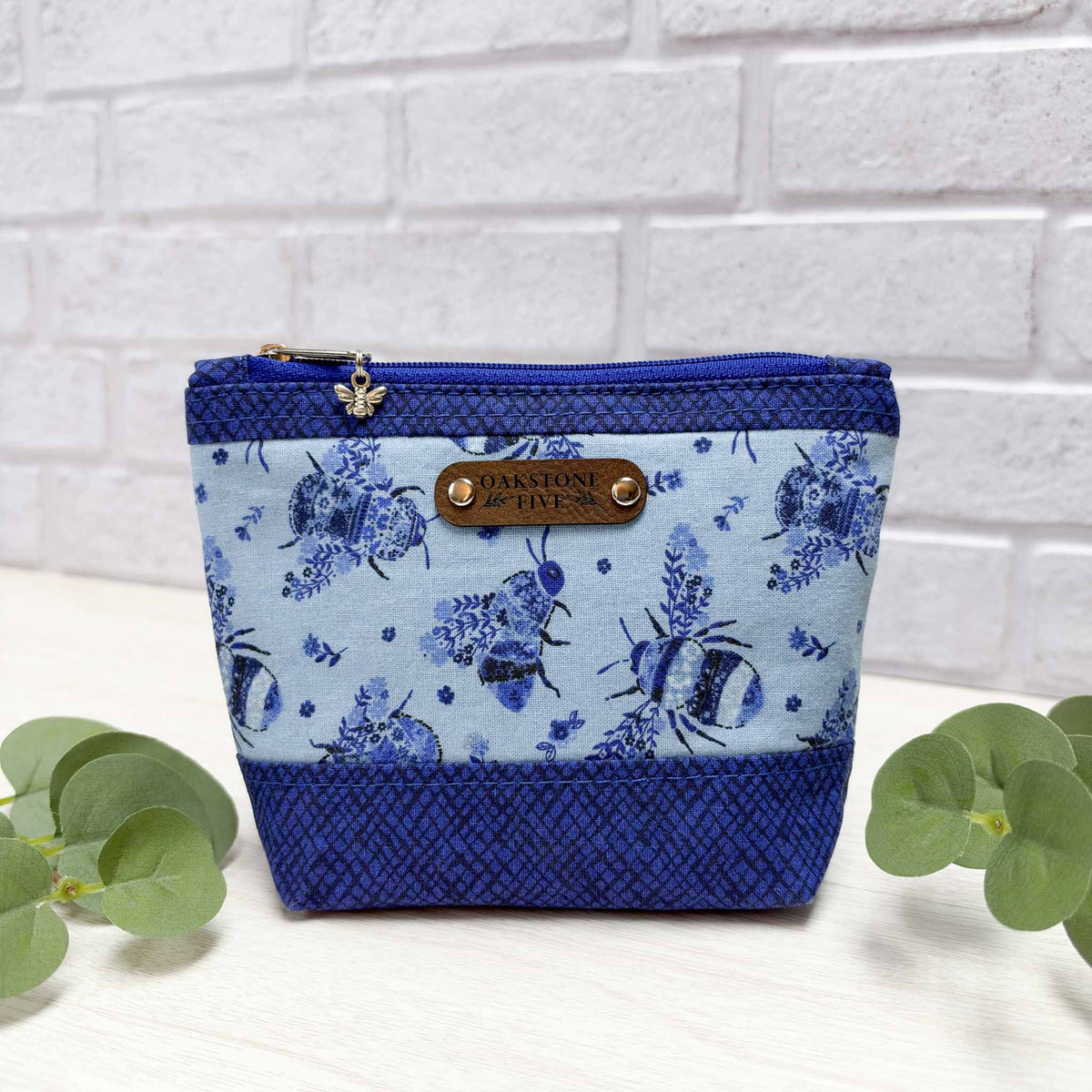 A blue small zipper pouch with a pollinator bee pattern accented with a royal blue fabric, sitting on a white surface with leaves, against a white brick wall.