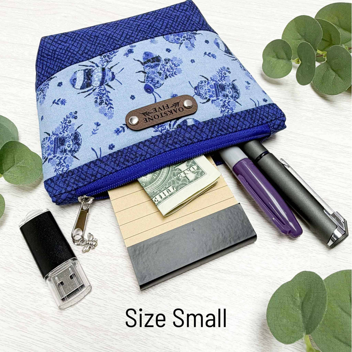 A cute flowers and bees zipper pouch, laying on a white surface with leaves and has pens and stationery items coming out of it.