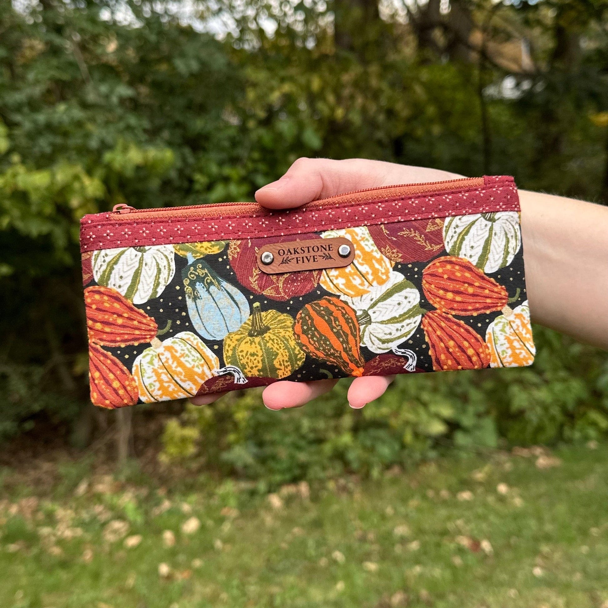 Hand holding a small zipper pouch with a Fall pumpkin and gourd pattern against a blurred green outdoor background