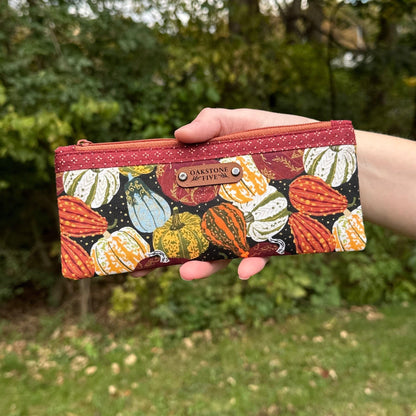 Hand holding a small zipper pouch with a Fall pumpkin and gourd pattern against a blurred green outdoor background