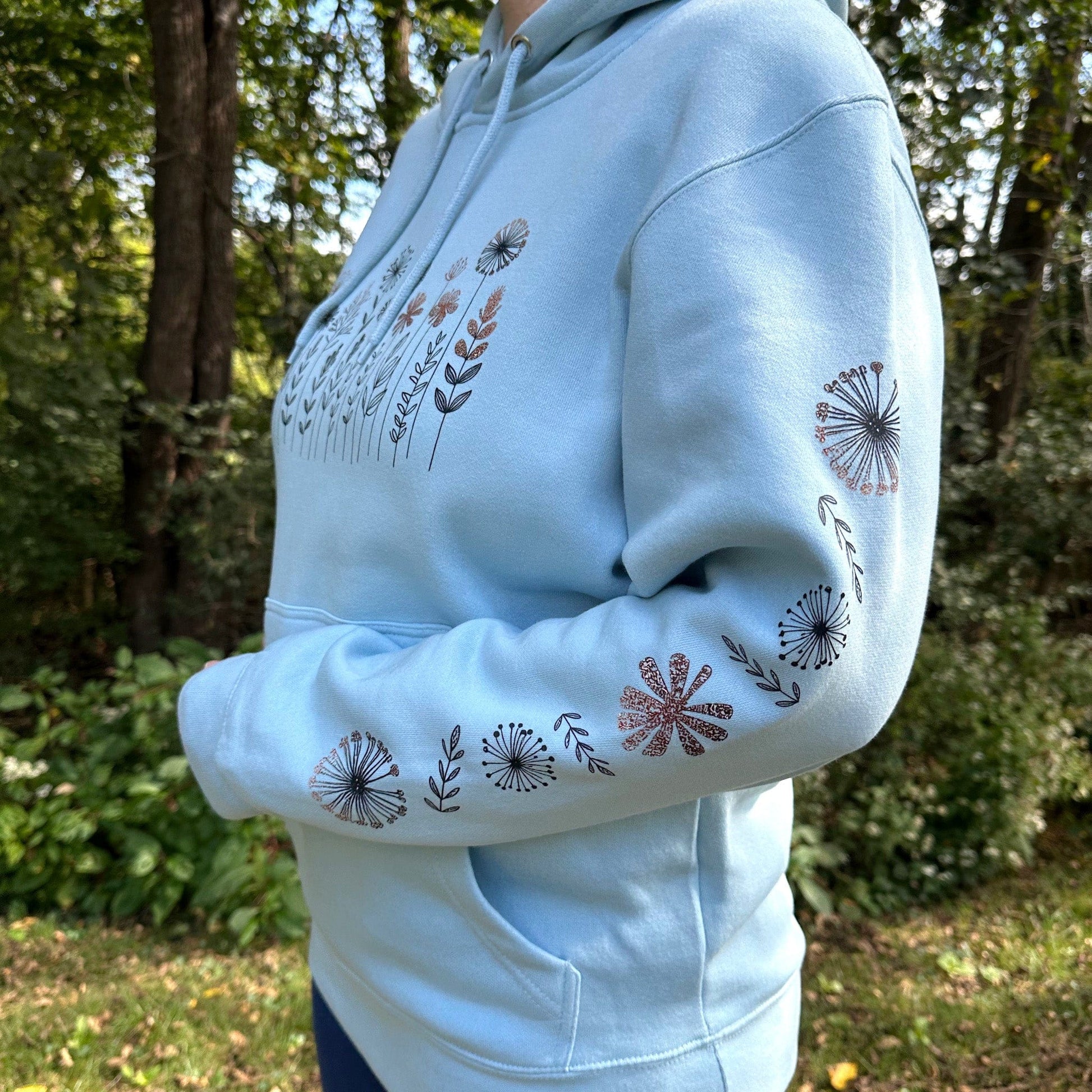 Woman standing in the woods wearing a light blue hoodie that has a flower design on the chest and down the sleeve.