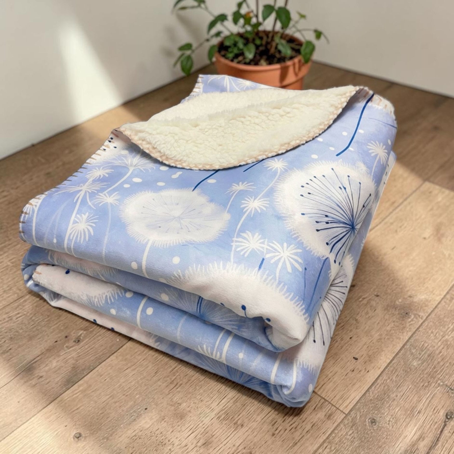 Dandelion patterned sherpa blanket laying folded on a table with a potted plant
