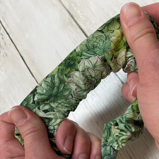 A close-up of a green, handmade scrunchie with a plant succulent pattern laying on a wooden surface
