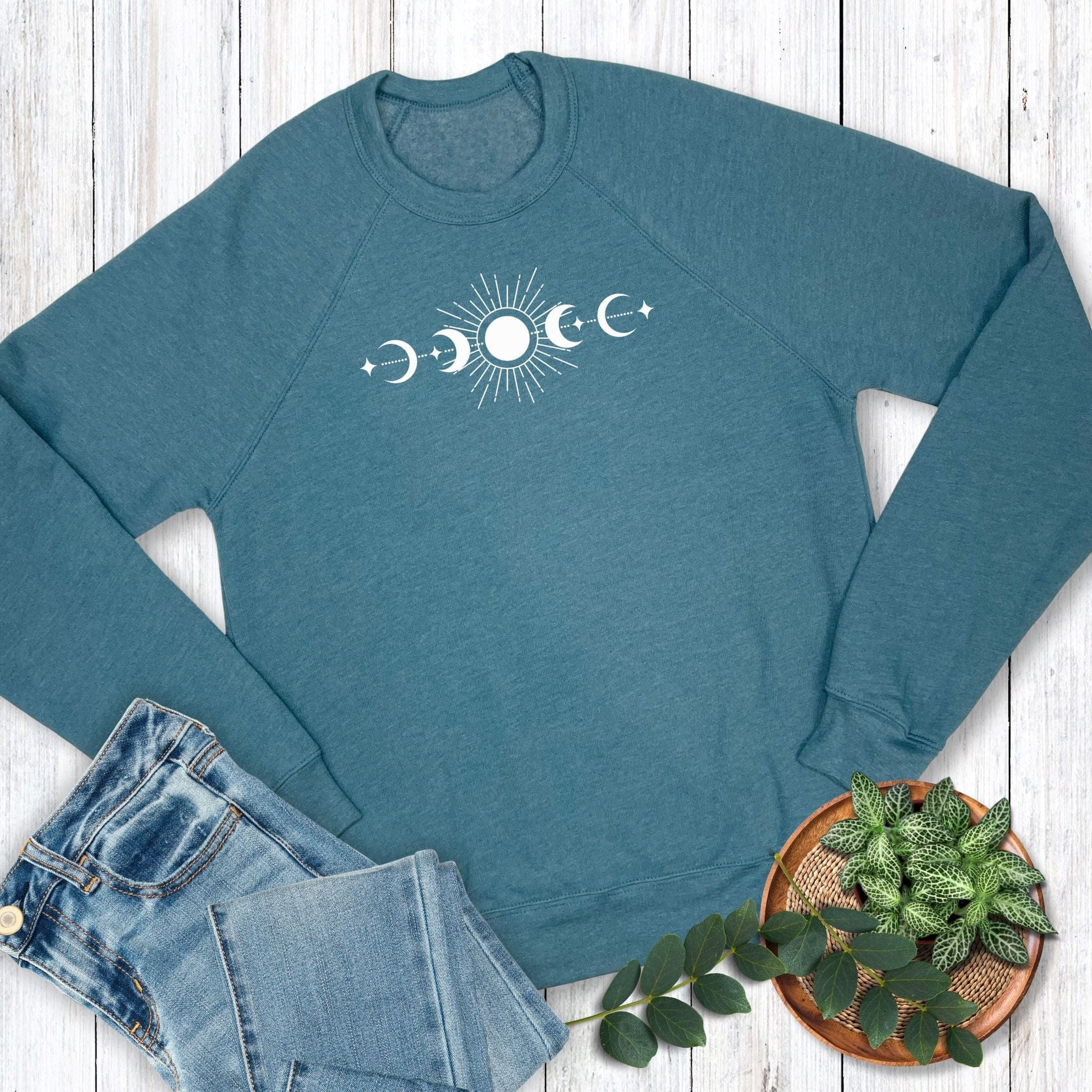 Teal sweatshirt with moon phase design on a wooden surface with jeans and a plant.
