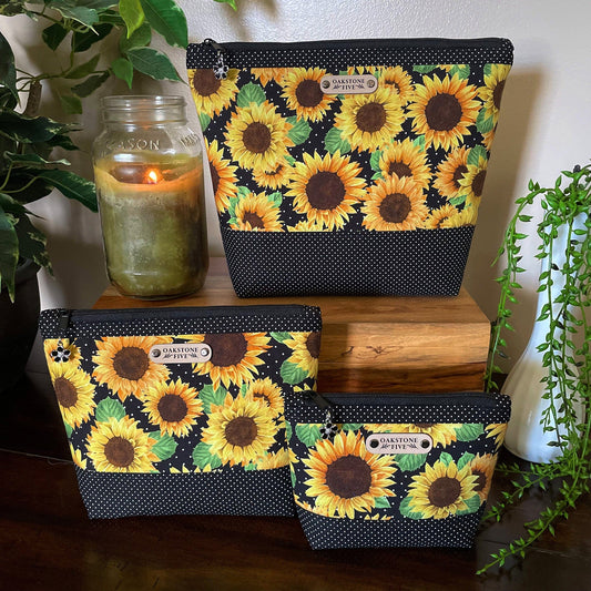 Set of three sunflower-patterned bags on a wooden surface with plants and a candle in the background.