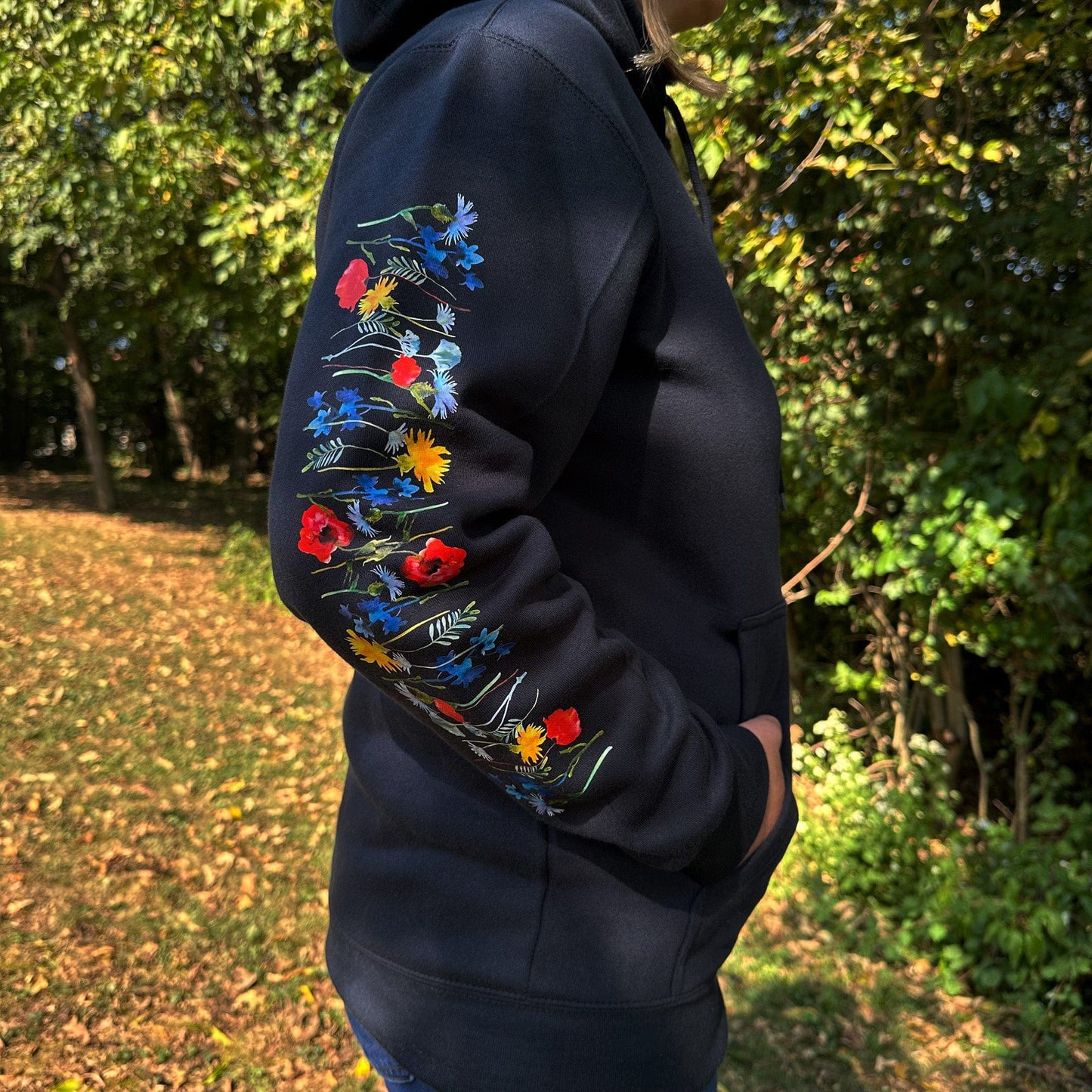 Woman standing outside in the woods, wearing a navy hoodie with vibrant floral designs on the sleeves.