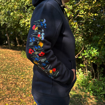 Woman standing outside in the woods, wearing a navy hoodie with vibrant floral designs on the sleeves.