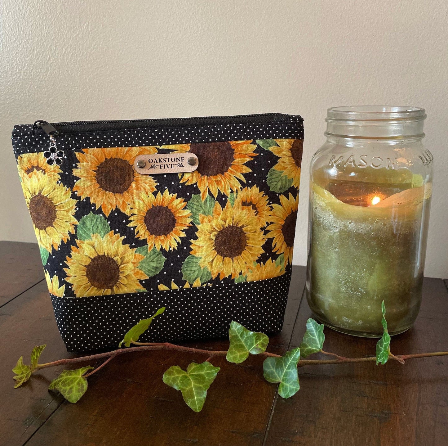 A medium sized sunflower zipper pouch on a wooden surface with a candle and ivy