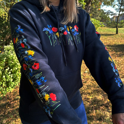 Woman standing outdoors, wearing a navy hoodie with floral designs on the sleeves and chest.