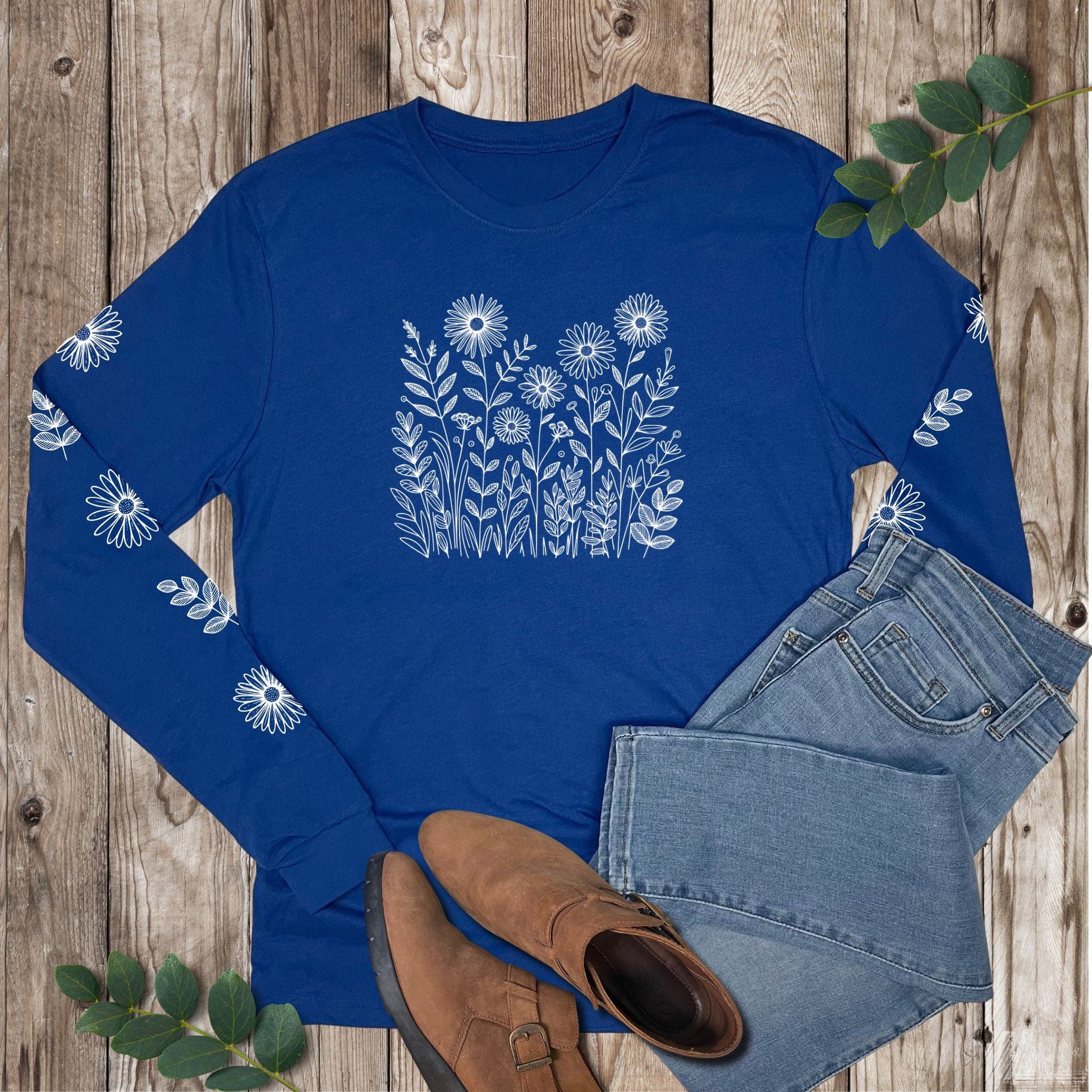 Blue long-sleeve shirt with white floral design on the front and sleeves, laying on a wooden surface with jeans and boots.