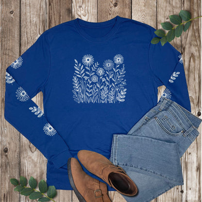 Blue long-sleeve shirt with white floral design on the front and sleeves, laying on a wooden surface with jeans and boots.