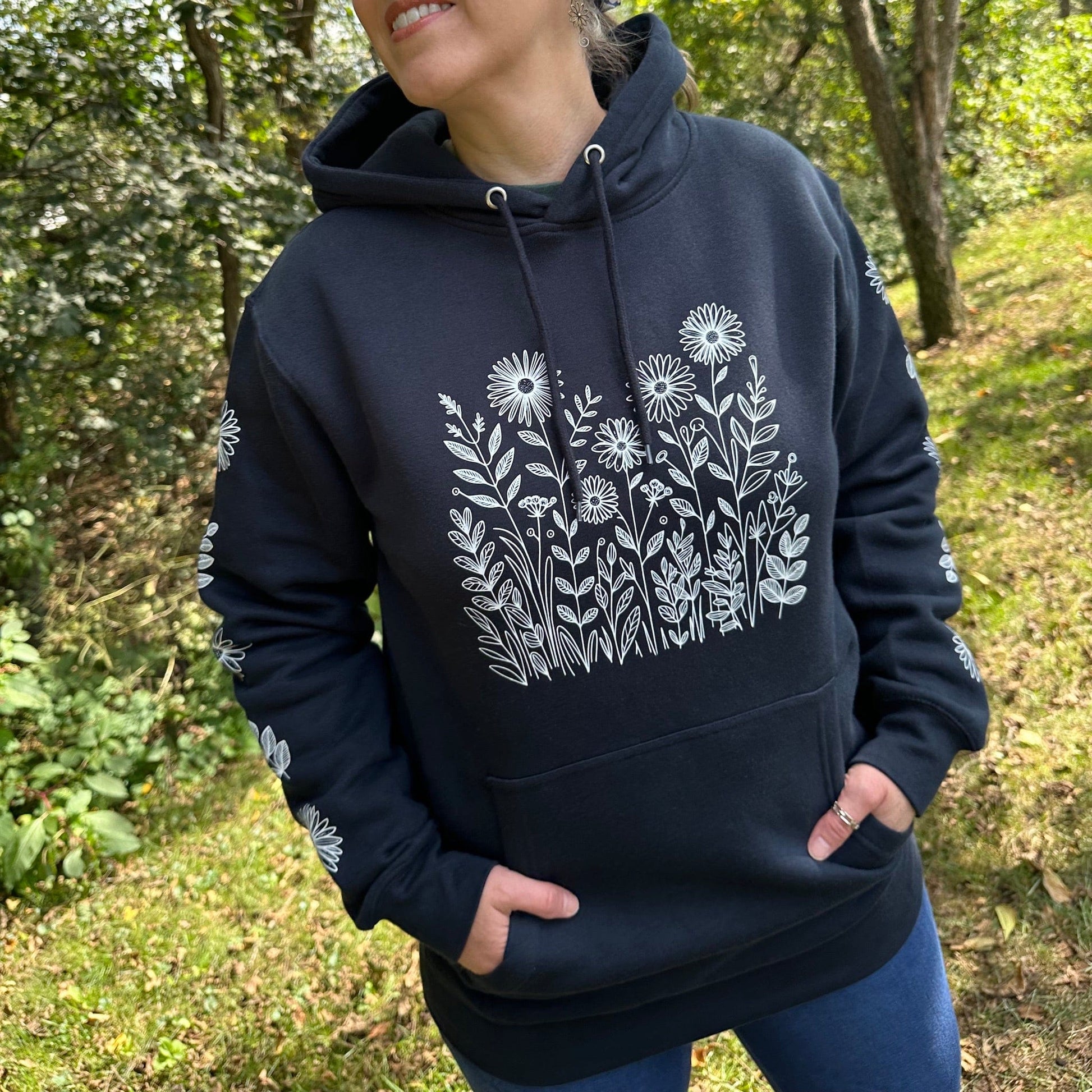 Woman standing outside in the woods, wearing a navy hoodie with a white flower design on the front and down the sleeves.
