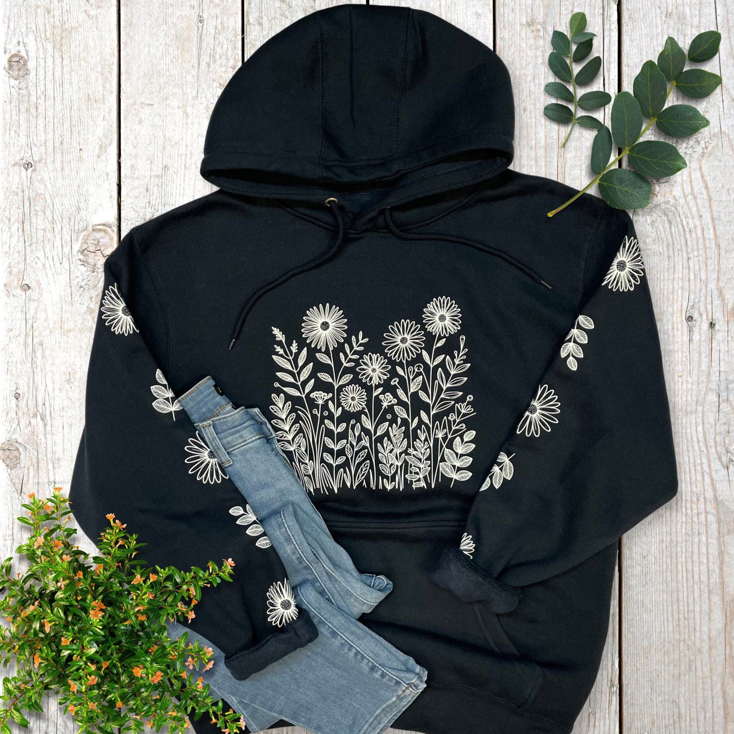Navy hoodie with white flowers on the front and sleeves,  laying on a wooden surface with denim jeans and greenery