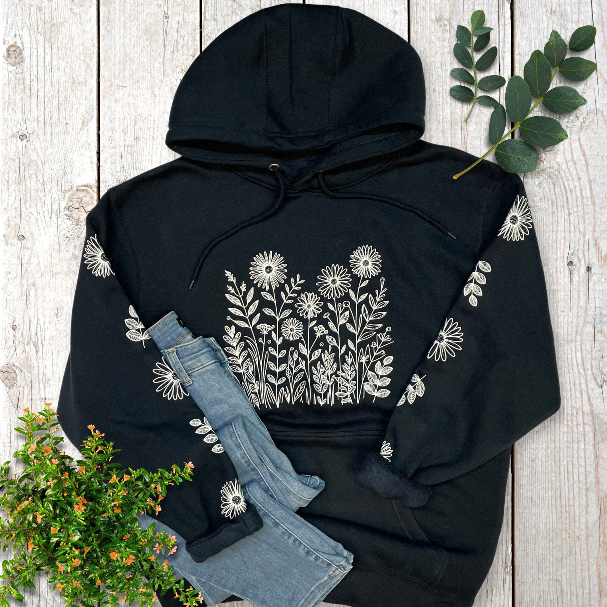 Navy hoodie with white flowers on the front and sleeves,  laying on a wooden surface with denim jeans and greenery
