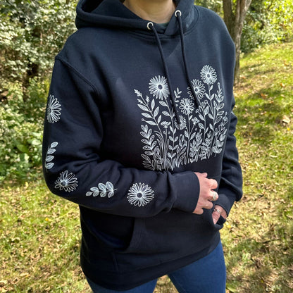 Woman standing outside in the woods, wearing a navy hoodie with a white flower design on the front and down the sleeves.